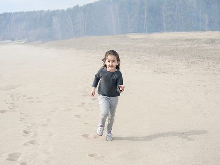 Little smiling sweet girl with brown hair running and jumping on the beach. Vacation by the sea. Cute kid girl on the deserted beach. Summer, outdoors. Wind in the hair girl. Beach landscape, desertの写真素材