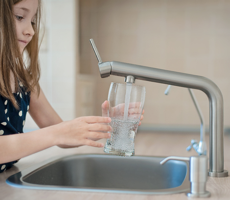 Portrait of a little caucasian girl gaining a glass of tap clean water. Kitchen faucet. Cute curly kid pouring fresh water from filter tap. Indoors. Healthy life conceptの写真素材