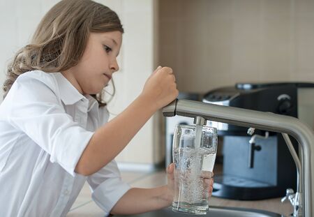 Portrait of a little caucasian girl gaining a glass of tap clean water. Kitchen faucet. Cute curly kid pouring fresh water from filter tap. Indoors. Healthy life conceptの写真素材