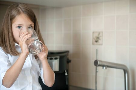 Portrait of a little caucasian baby girl holding a glass of tap clean water. Kitchen faucet. Cute kid pouring fresh water from filter tap. Indoors. Healthy life conceptの写真素材