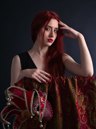 Beautiful redheaded woman in a black dress with carnival maskの写真素材