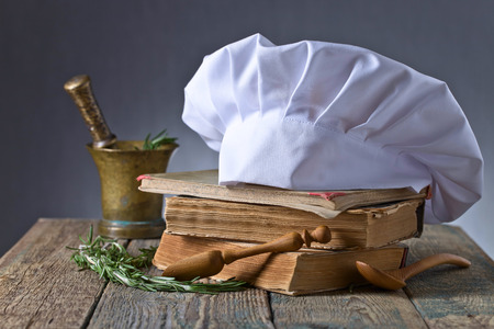 Old copper mortar with rosemary. Culinary books , chef hat and wooden spoons . Kitchen accessories on the old wooden table .