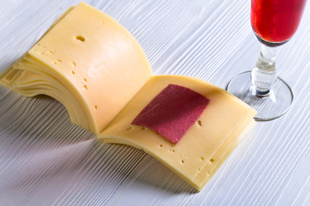 Slices of cheese folded like a book and glass of red wine on a white wooden table .の写真素材