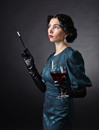 Portrait of young  woman in retro style with cigarette and glass of red wine. Beautiful brunette with perfect hairstyle and  professional  make-up in retro stile . Long evening dress.の写真素材