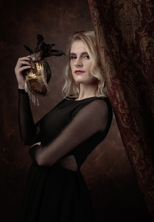 Portrait of beautiful young woman with carnival mask. Gorgeous hair and natural makeup. Fashion beauty photo. Natural blonde in black dress.の写真素材