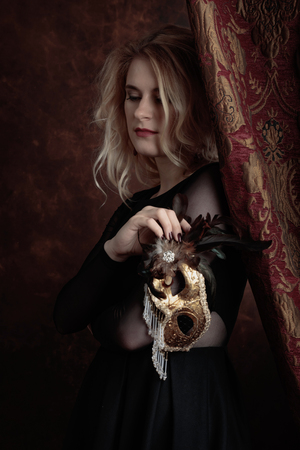 Portrait of beautiful young woman with carnival mask. Gorgeous hair and natural makeup. Fashion beauty photo. Natural blonde in black dress.の写真素材