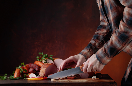 Man in a brown plaid shirt cuts various sausages and smoked meat on an old wooden table . Copy space .の写真素材
