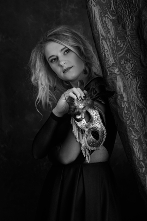 Portrait of beautiful young woman with carnival mask. Gorgeous hair and natural makeup. Fashion beauty photo. Natural blonde in black dress.の写真素材