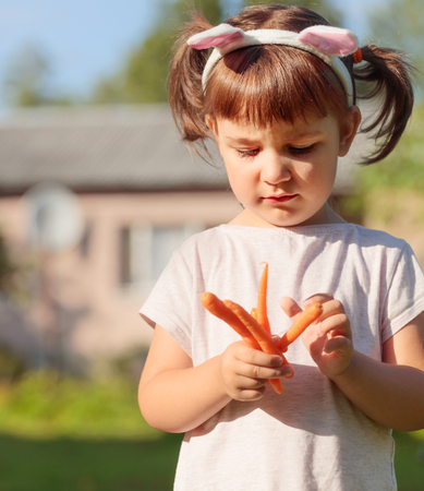 Happy little girl eating fresh and sweet carrot. European little girl with rabbit ears on a Sunny summer day in the garden.の写真素材
