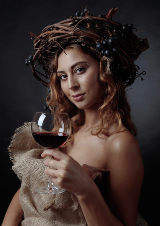 Beautiful girl with long wavy hair and perfect make-up. Glass of red wine in hand and vine wreath with blue grapes on a head.の写真素材