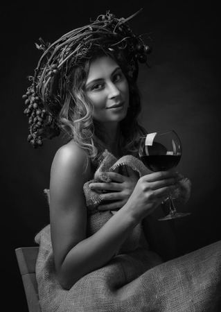 Beautiful girl with long wavy hair and perfect make-up. Glass of red wine in hand and vine wreath with blue grapes on a head, black and white.の写真素材