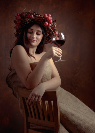 Beautiful girl with long wavy hair and perfect make-up. Glass of red wine in hand and vine wreath with grapes on a head.の写真素材