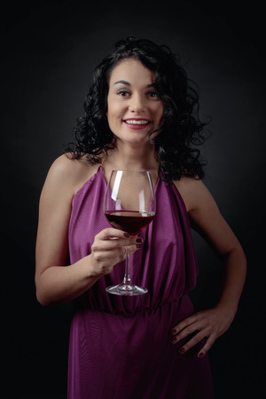 Young beautiful woman with long curly hair in  purple evening gown. Portrait of attractive brunette in long evening dress with glass of red wine.の写真素材