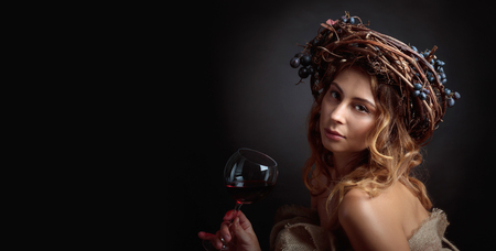 Beautiful girl with long wavy hair and perfect make-up. Glass of red wine in hand and vine wreath with blue grapes on a head. Copy space for your text.の写真素材