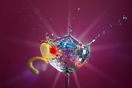 Colorful cocktail in glass with splashes garnished with lemon and cherries. Purple background. Mixed light. Copy space for your text.の写真素材