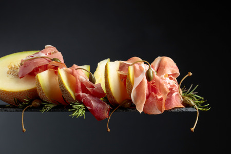 Prosciutto with melon, rosemary and capers on a black background, copy space.の写真素材