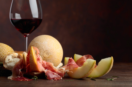 Glass of red wine and mediterranean snacks. Prosciutto with melon and rosemary on a old wooden table, copy space.の写真素材