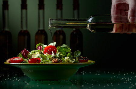 Green salad with cherry tomatoes, sesame and walnuts watered with vegetable oil. In the background, silhouettes of olive oil bottles.の写真素材
