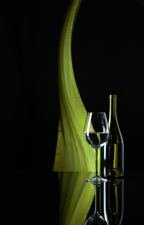 Wineglass and bottle of white wine on a black reflective background. Green satin curtain flutters in the wind. Copy space.の写真素材