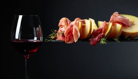 Glass of red wine and prosciutto with melon, capers and rosemary on a black background, copy space.の写真素材