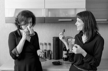 Two middle-aged girlfriends talk and drink coffee in the kitchen. Black and white.の写真素材
