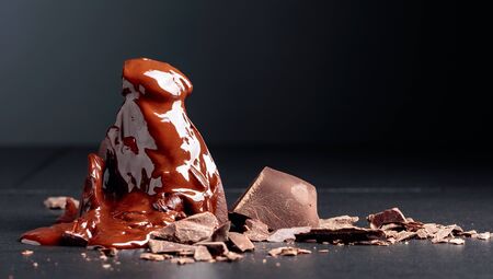 Broken large piece of dark chocolate with crumbs on a black table. Large piece melts and drips onto...