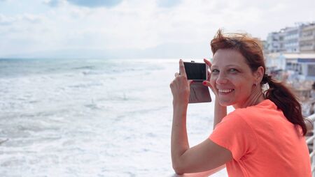 Attractive mature traveler woman use mobile phone take a photo of beautiful seascape. Copy space.の写真素材