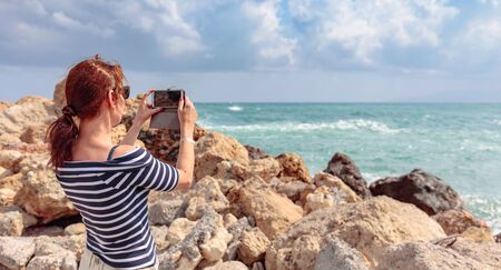 Attractive mature traveler woman use mobile phone take a photo of beautiful seascape. Copy space.の写真素材