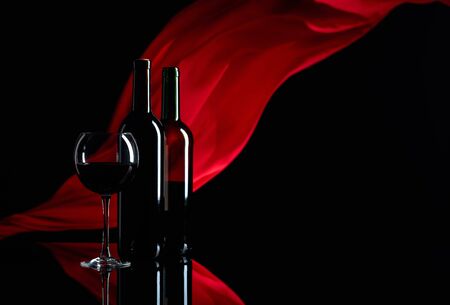 Wineglass and bottles of red wine on a black reflective background. Red satin curtain flutters in the wind. Focus on foreground. Copy space.の写真素材