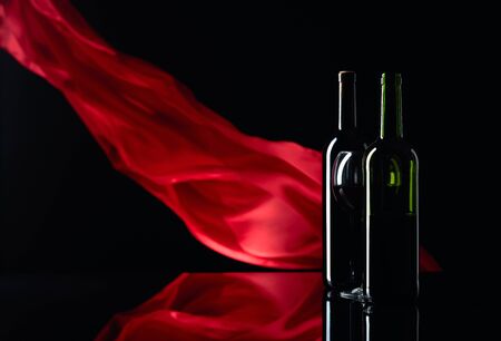 Wineglass and bottles of red wine on a black reflective background. Red satin curtain flutters in the wind. Focus on foreground. Copy space.の写真素材