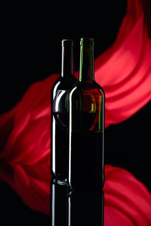 Wineglass and bottles of red wine on a black reflective background. Red satin curtain flutters in the wind. Focus on foreground. Copy space.の写真素材