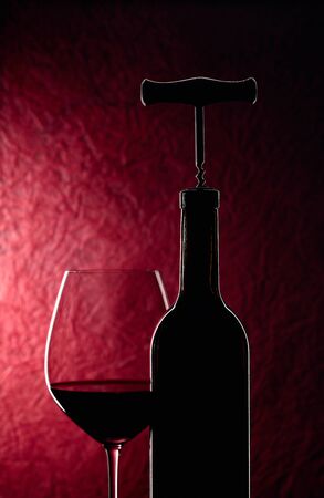 Bottle with corkscrew and glass of red wine on a dark red background.の写真素材