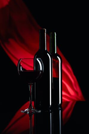 Wineglass and bottles of red wine on a black reflective background. Red satin curtain flutters in the wind. Focus on foreground. Copy space.の写真素材