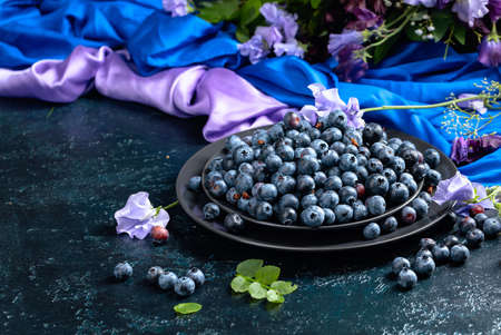 Fresh juicy blueberries on a black plate. Summer still life with blueberries, colored sweet peas and meadow grasses on a dark blue background.の写真素材