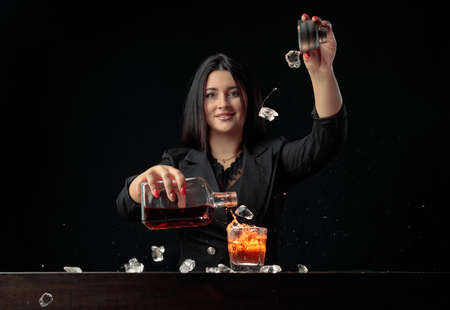 Attractive bartender girl pours an alcoholic drink into a glass and throws pieces of ice. Beautiful brunette in black prepare cocktail.の写真素材