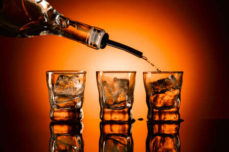 Strong alcohol is poured into glasses with ice. Black reflective background and orange backlit. Copy...