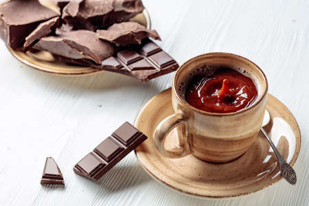 Hot chocolate and broken pieces of bitter chocolate on a white wooden table. A drop of drink falls...