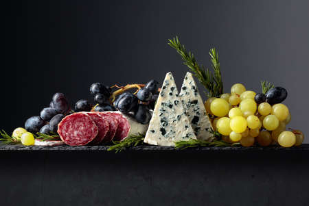 Blue cheese, dry-cured sausage, grapes, and rosemary on a black background. Simple and tasty food....