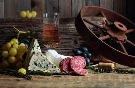 Rustic still life with wine and snacks. On an old wooden table wine, blue cheese, dry-cured sausage,...