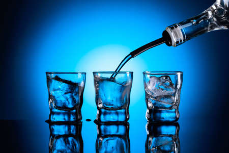 Vodka is poured from bottle into glasses with ice. Frozen glasses on a dark blue background. Copy space.の写真素材