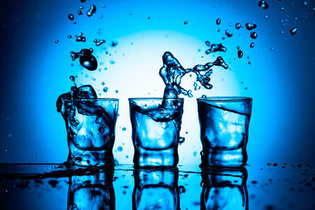 Strong alcoholic drink in dammed glasses with ice on a black reflective background. Splashes on a blue background.の写真素材