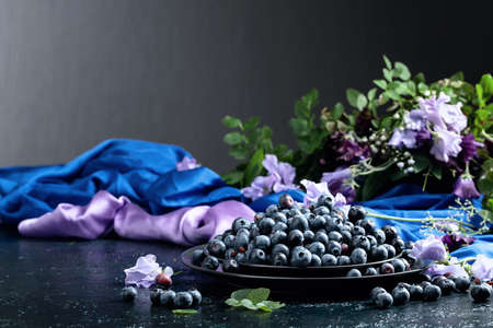 Fresh juicy blueberries on a black plate. Summer still life with blueberries, colored sweet peas and meadow grasses on a dark blue background.の写真素材