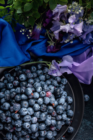 Fresh juicy blueberries on a black plate. Summer still life with blueberries, colored sweet peas and meadow grasses on a dark blue background. View from the top.の写真素材