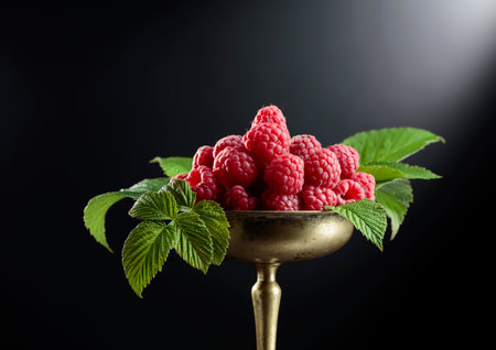 Fresh raspberries with leaves in an old brass dish on a black background.