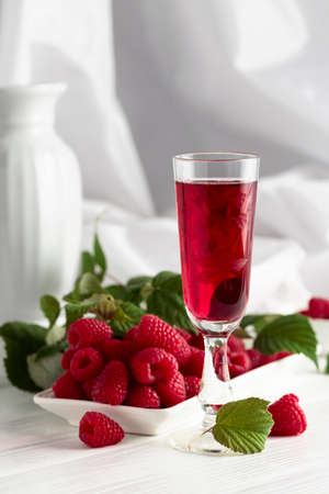 Raspberry liquor and fresh berries with leaves on a white wooden table.
