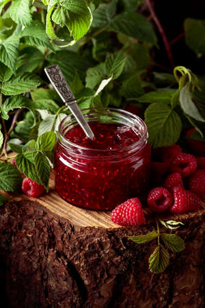 Jar of raspberry jam and fresh berries with leaves on a pine stump in the forest.