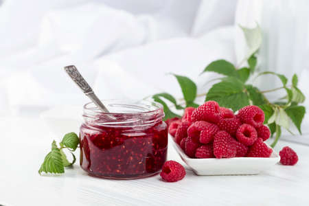 Jar of raspberry jam and fresh berries with leaves on a white wooden table.の写真素材