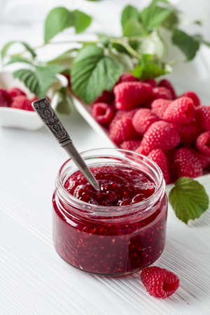 Jar of raspberry jam and fresh berries with leaves on a white wooden table.の写真素材