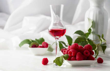 Raspberry liquor and fresh berries with leaves on a white wooden table.