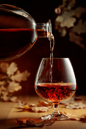 Cognac is poured into a glass from a bottle. A snifter of brandy on an old oak table with fallen oak...